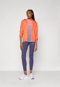 Tenue de sport comprenant une veste à capuche légère orange, des leggings violets et des chaussures de course violettes claires avec des détails blancs.