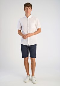 Camisa blanca de manga corta con botones y bolsillo en el pecho, combinada con pantalones cortos azul marino arremangados y zapatillas blancas. Diseño simple y casual.