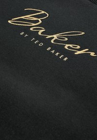 Melnā auduma ar zeltījuma izšuvumu, kurā ir uzraksts "Baker by Ted Baker". Struktūra izskatās mīksta un gluda, ar klasisku, minimālistisku dizainu.
