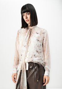Eine Frau mit glattem, dunklem Haar trägt eine durchsichtige, hellfarbige Bluse mit einer langen Krawatte und einer dunkelbraunen Lederhose, während sie vor einem weißen Hintergrund steht.