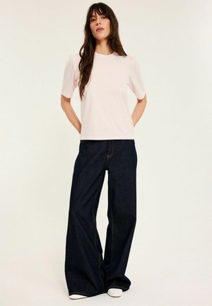 Kobieta o długich brązowych włosach, ubrana w jasnoróżowy top z krótkim rękawem i ciemne, szerokie dżinsy, stojąca na jednolitym tle.