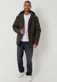 Mörkgrön isolerad jacka med huva, dragkedja fram och sidofickor, buren över en vit t-shirt och kombinerad med gråbyxor och vita sneakers.