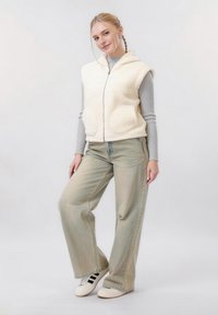 Jeune femme aux cheveux tressés portant un gilet en polaire blanc, un haut à manches longues côtelé gris, un jean à jambes larges et des baskets blanches, sur un fond uni.