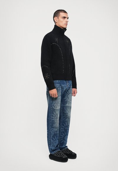 Črna zip-up jopica s zakovicami. Nosi se z vzorčenimi modrimi kavbojkami in črnimi slip-on čevlji. Model stoji v profilu pred belo ozadjem.