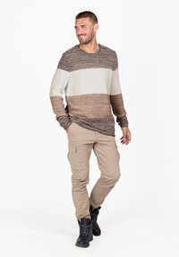 Maglione a righe in tonalità beige e marrone, abbinato a pantaloni cargo khaki e stivali neri con lacci, in piedi su uno sfondo bianco.