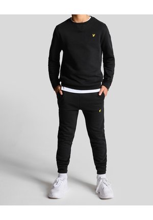 Personne portant un sweat-shirt noir et un jogging assorti avec des logos jaunes, un t-shirt blanc en dessous, des chaussettes blanches et des baskets blanches, debout les mains dans les poches.