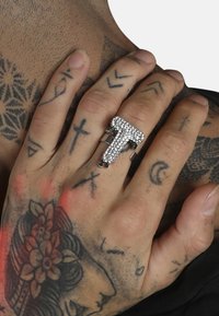 Anillo plateado con forma de la letra "T", adornado con piedras de strass transparentes y acentos verdes, llevado en una mano tatuada.