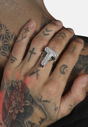 Anillo plateado con forma de la letra "T", adornado con piedras de strass transparentes y acentos verdes, llevado en una mano tatuada.