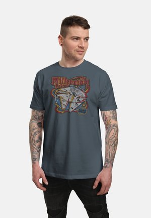 Mężczyzna z tatuażami, ubrany w ciemnoniebieską koszulkę z nadrukiem Gwiezdnych Wojen przedstawiającą Sokoła Millennium oraz czarne podarte dżinsy, patrzący nieco w prawo.