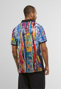Bărbat cu păr scurt și tatuaj, purtând o cămașă cu mânecă scurtă, colorată și cu modele, cu guler negru și cuvântul "WAVEBUHEL" pe spate.