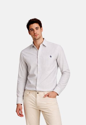 Jeune homme aux cheveux courts et foncés portant une chemise à manches longues rayée gris clair et un pantalon beige, debout avec une main dans la poche.