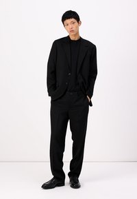 Veste de costume noire avec revers crantés, associée à un shirt noir à col rond et un pantalon ample. Porté avec des chaussures noires cirées.