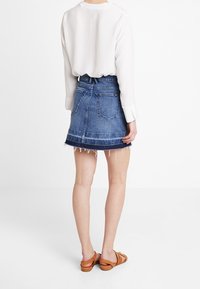 Jeanskjol med fransad nederkant, blå färg och två bakfickor. Bärs med en lös, vit långärmad topp och bruna sandaler.