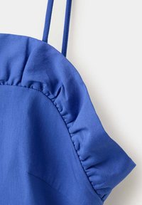 Robe en tissu bleu avec fines bretelles et détail à volants le long de la poitrine et du bord des manches sur fond blanc.