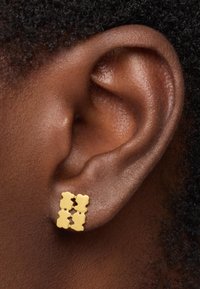 Złote kolczyki z projektem czterech połączonych kształtów niedźwiedzi, osadzone na teksturowanym tle w odcieniu skóry w zbliżeniu.