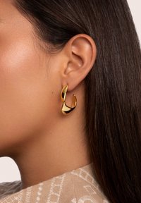 Pendiente de aro abstracto en tono dorado con un acabado liso y brillante. Presenta un diseño en forma de ola, adherido al lóbulo de la oreja.