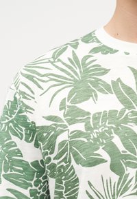 Άσπρο πουκάμισο με πράσινο τροπικό σχέδιο φύλλων που φοριέται από άτομο, εμφανίζοντας τον ώμο και μέρος του λαιμού σε απλό φόντο.