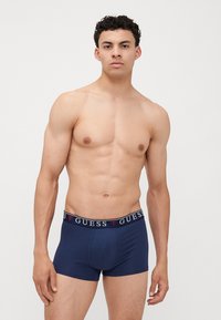 Boxeri scurți de culoare bleu marin, cu un brâu elastic branduit pe care este inscripționat "GUESS" cu litere albe. Textură netedă și design strâns.