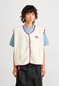 Cream fleece vest with a zip front, maroon trims, and two side pockets, layered over a blue t-shirt, featuring a logo patch on the chest.