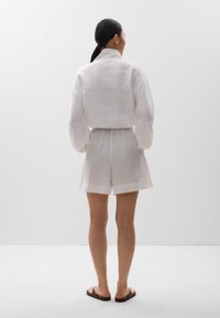 Weißes Leinenhemd und Shorts-Set mit gerafftem Bund, ausgestattet mit langen Ärmeln und einem Kragen. Flache Sandalen runden das Outfit ab.