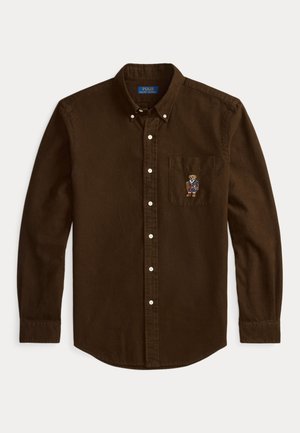 Camisa castanha de botão com mangas longas, em material de algodão, apresentando um bolso no peito com um design de urso bordado. Gola clássica e botões brancos.