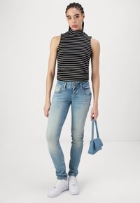 Top sin mangas a rayas en blanco y negro, jeans ajustados azules, zapatillas blancas, sosteniendo un pequeño bolso azul; atuendo casual con texturas variadas.
