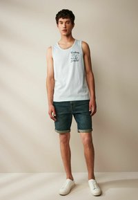 Ljusblå tanktopp med grafisk tryck, tillsammans med mörka denimshorts uppvikta vid fållen. Vita sneakers fulländar den avslappnade looken.