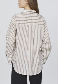 Stribet langærmet skjorte i creme og brun, med knap-ned krave, løs pasform og buet nederkant, samt opdrejede ærmer.