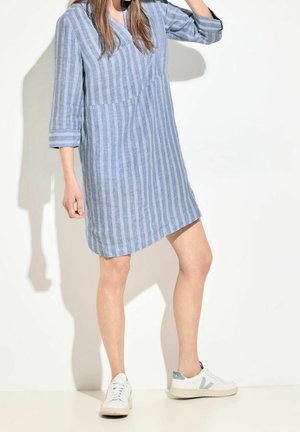Femme portant une robe à rayures bleues et blanches longueur genou avec des manches 3/4, associée à des baskets blanches et grises, debout contre un mur blanc.