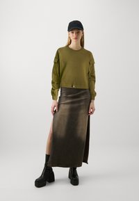 Sudadera corta de color verde oliva con mangas abullonadas, combinada con una falda maxi de corte alto en marrón oscuro. Una gorra negra y botas de tacón grueso completan el look.