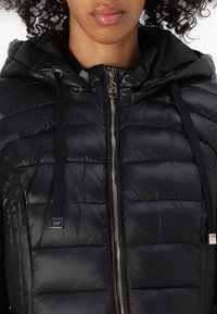 Zwarte pufferjas met een capuchon, voorzien van een ritsluiting, voelbare structuur en zwarte trekkoorden. Inclusief een klein zilveren logo-tag.