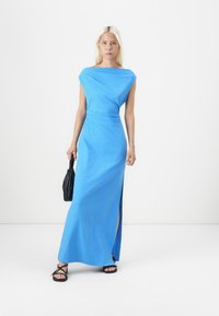 Μπλε, φόρεμα μέχρι τον πάτωμα με σχεδίαση off-shoulder, πλευρική σχισμή και λεία υφή. Φοριέται με μια μαύρη τσάντα χεριού και ελάχιστα σανδάλια.