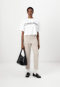 Hvit cropped t-skjorte med "Calvin Klein" logo, beige skreddersydde bukser, svart lærveske, og svarte loafers med hvite sokker.