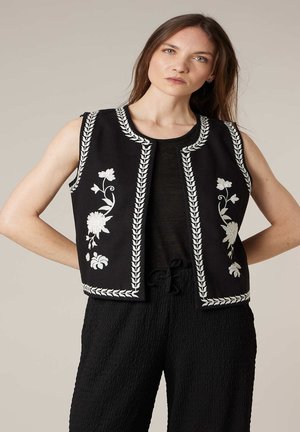 Femme portant un gilet noir sans manches avec une broderie florale blanche sur un haut noir et un pantalon noir texturé, debout les mains sur les hanches.