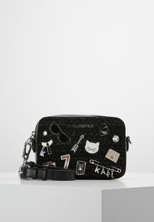 Zwarte glitter schoudertas met zilveren ketting, leren riem en diverse metalen en emaille speldjes, waaronder een kat, een zonnebril, een pijl en tekstbedeltjes.