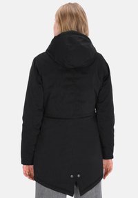 Person med skulderlangt blondt hår iført en sort hættetrøje med fishtail-kant og to metalknapper på bagsiden.