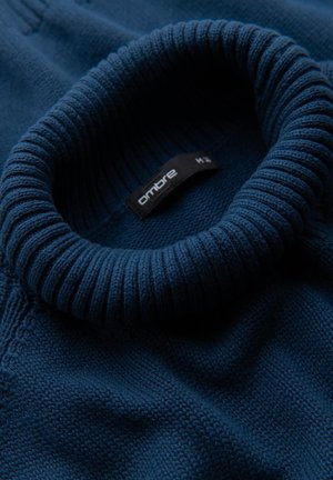 Navy gebreid sweater met een geribde hoge kraag. De stof heeft een gestructureerd patroon en een label met de tekst "ombre M." in close-up weergave.