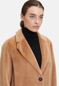 Cappotto in pelliccia sintetica beige con revers, chiusura a un bottone nero, e texture morbida, indossato sopra un maglione a collo alto nero.