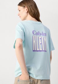 Lichtblauwe katoenen t-shirt met een groot paarse "Calvin KLEIN" opdruk op de achterkant. Korte mouwen, relaxte pasvorm, zachte textuur.