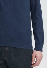 Maglione lavorato a maglia blu navy con maniche lunghe, polsini e orlo a costine, con etichetta tessuta sul lato vicino al fondo.