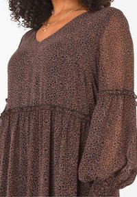 Mujer que lleva un vestido marrón con estampado de leopardo, con largas mangas de tul abullonadas y escote en V, complementado con collares de oro en capas.