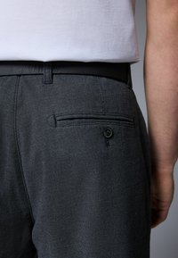 Pantalons gris texturé avec une poche arrière dotée d'un bouton noir, portés avec une chemise blanche unie. Mettez l'accent sur les détails du tissu et de la couture.