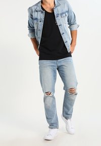 Mężczyzna w jasnoniebieskich podartych dżinsach, czarnej koszulce, jasnoniebieskiej kurtce dżinsowej z podwiniętymi rękawami i białych trampkach idący do przodu.