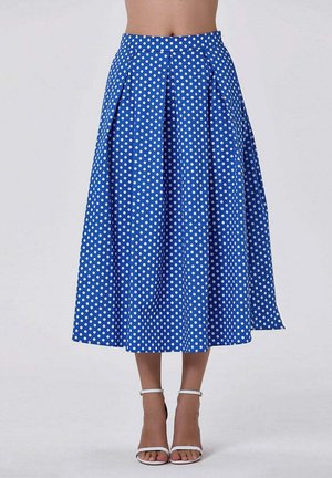 Jupe midi bleue taille haute à pois blancs, plissée, portée avec des sandales à talons hauts blanches à bride à la cheville, montrée de la taille aux pieds.
