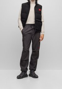 Schwarzige gepolsterte Weste mit einem roten Logo, cremefarbener Rollkragenpullover, graue Cargo-Hosen mit Taschen und schwarze Sportschuhe mit roten Akzenten.
