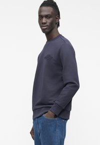 Tmavě modrá mikina s kulatým výstřihem a žebrovanými manžetami. Na hrudi vyšitá logo. Kombinováno s modrými džíny.