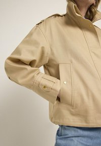 Beige jacket with a high collar, shoulder epaulettes, and a front pocket. Features a relaxed fit and gold snap button accents.