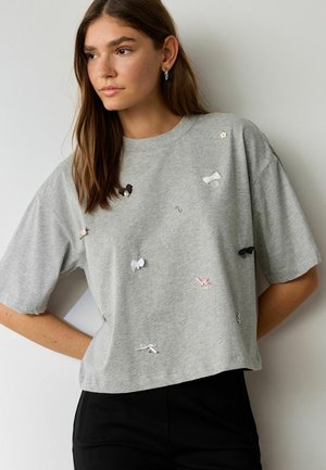 Tânără cu păr lung și castaniu, purtând un tricou gri larg decorat cu mici funde și ace de cusut, asortat cu pantaloni negri.