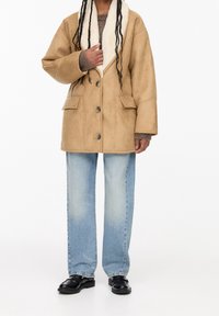 Tan faux læder frakke med en hvid pelskrave, oversized pasform, tre knapper og to frontlommer, båret over lyseblå jeans og sorte sko.