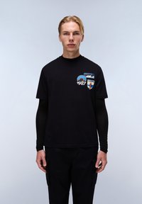 Sort kortærmet t-shirt med bjerg- og skjoldgrafik i blå, hvid og orange farver; båret over lange sorte ærmer.